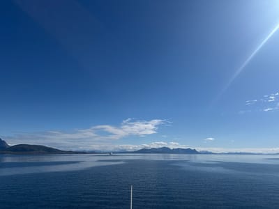A wide-angle, high-angle shot captures a serene seascape under a bright, clear blue sky with a prominent sunbeam extending from the upper right corner. The foreground is dominated by the deep blue, rippling surface of the water, with a single white pole extending vertically from the bottom center. In the mid-ground, a distant white ferry is visible sailing across the water. The background features a chain of majestic, dark, and somewhat rugged mountains, some with snow-capped peaks, receding into the distance. The coastline is dotted with islands and fjords, typical of the Norwegian landscape. Wispy white clouds are scattered across the sky, adding depth and texture to the scene. The overall impression is one of tranquility and natural beauty. The scene is consistent with views from the town of Lødingen, Norway, known for its stunning fjord and mountain scenery.