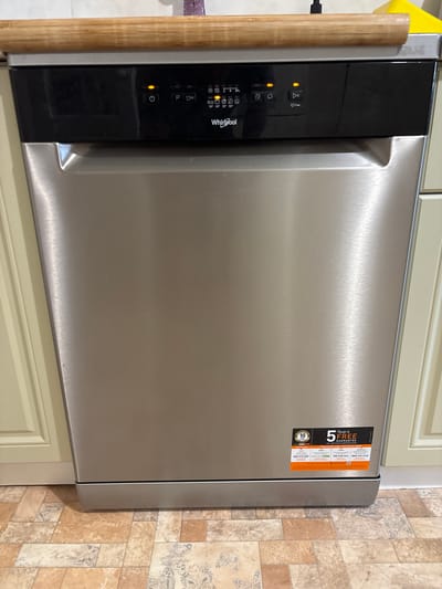 An eye-level, medium shot shows the top portion of a stainless steel dishwasher. The dishwasher is integrated into a kitchen with cream-colored cabinets. Above the dishwasher is a light-colored wooden countertop. The dishwasher has a black control panel with several illuminated buttons and indicators, including a power button, a "P" button, and icons for wash cycles and temperatures. The Whirlpool logo is visible on the control panel. Below the control panel, the stainless steel front of the dishwasher is clean and reflects light. A sticker on the lower right of the dishwasher front displays "10 Years GUARANTEE" and "5 Years FREE GUARANTEE REGISTER AND ACTIVATE NOW". The floor in front of the dishwasher has a tiled pattern with beige and brown squares, some of which have a faint butterfly-like design. The overall scene is indoors, in what appears to be a domestic kitchen. The location is the city of Pohreby, Ukraine.