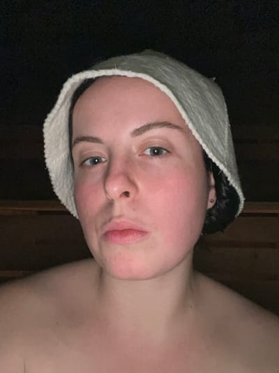 A person with fair skin, blue eyes, and a neutral expression wears a white wool hat. The person's cheeks are flushed with red, and their hair is dark. The subject appears to be in an indoor setting, possibly a sauna. Dark wood paneling is faintly visible in the background. The person's shoulder is bare.