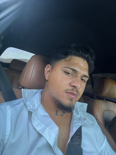 In an indoor setting that appears to be the interior of a vehicle, a young man of evident Mediterranean or Middle Eastern descent is captured in a close-up, angled selfie. He is seated, with the car's dark interior and brown leather seats visible in the background, suggesting a vehicle with a premium finish. The man has dark, wavy hair styled upwards, and a noticeable amount of facial hair, including a mustache and a goatee, along with what appears to be acne on his forehead and cheeks. He is wearing a white dress shirt, open at the collar, revealing a black seatbelt across his chest and intricate tattoos on his neck and upper chest, one of which prominently displays the number "12". The lighting suggests a daytime setting, possibly with natural light filtering into the car. The image does not provide any specific information or visual cues to identify the location as Dolna Vasilitsa, Bulgaria.