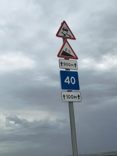 This image shows a traffic signpost with multiple signs. The signs are stacked vertically on a metal pole. The top sign is a triangular warning sign depicting a car skidding, indicating a slippery road. Below that is another triangular warning sign showing a downward slope with "6%" written on it, signifying a steep descent. Following these are three rectangular signs. The first indicates "↑ 900m ↑", suggesting a distance of 900 meters ahead. The second, a blue sign with white numbers, displays "40", likely a speed limit. The final sign reads "↑ 100m ↑", indicating a distance of 100 meters. The signs are against a backdrop of a heavily clouded, overcast sky, suggesting a potentially stormy or gloomy weather condition. The lower part of the image shows a faint, blurred landscape, possibly indicating the location is on the outskirts of a city or in a more open area. The image was taken in Rogozhkin, Russia. There are no people visible in the image.