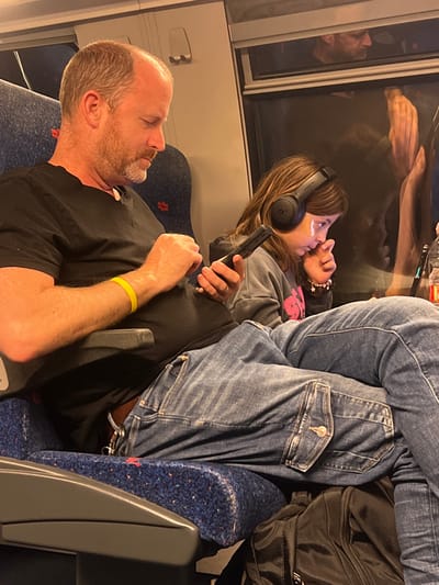 The image shows a man and a young girl sitting on a train. The man is wearing a black t-shirt, jeans, and a yellow wristband. He is using a smartphone with his right hand. The girl is wearing a grey sweatshirt and headphones. She is looking down at something in her lap, and using her left hand to cover her mouth. The girl is sitting on the seat in front of the man. The train has blue seats with speckled fabric. The man's right leg is stretched out in front of him. The girl's legs are tucked under the seat in front of her.  The man's left leg is bent and rests on the seat behind him. There are a few other people sitting in the window seats, but they are out of focus. There is a bag at the man's feet. The image is taken inside a train carriage. The coordinates 31.749, 34.823 place the image in the city of Kfar Menahem, Israel. The image suggests a normal, everyday scene of people commuting on a train.
