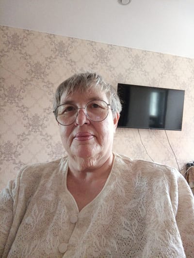 A medium-shot selfie of an elderly Caucasian woman with short, grey hair and glasses is taken indoors, in Gusinoozyorsk, Russia. She is wearing a light-colored, knitted cardigan over a white top. The background consists of patterned wallpaper and a flat-screen television mounted on the wall. The woman is looking directly at the camera, with her lips slightly parted. Her facial features are prominent, with visible wrinkles on her forehead and around her eyes. The lighting is bright and appears to be from an overhead source. The overall scene suggests a casual indoor setting, likely a living room.