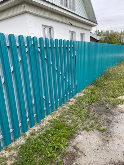The image depicts a turquoise metal fence running alongside a two-story house in Talmenka, Russia. The house has white walls, white trim, and windows. The fence runs along the edge of the property, separating the house from a dirt and gravel area overgrown with grass and weeds. In the background, lilac bushes are visible.