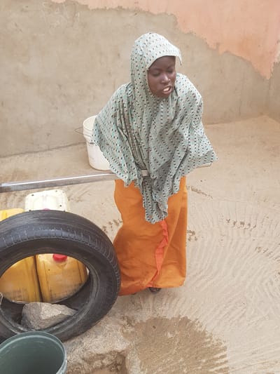 An image of a woman, likely from the city of Dutsinma A, Nigeria, wearing a patterned headscarf and an orange dress. She is seated on a white plastic chair. A tire labeled "MICHELIN" leans against containers. A green bucket is present, and the setting appears to be an outdoor area with a textured wall.