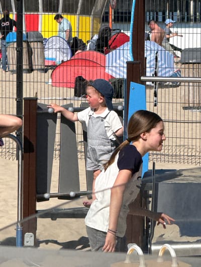 This image captures a vibrant, sunny outdoor scene, likely at a recreational area or boardwalk in Blankenberge, Belgium. The photo appears rotated, showing a dynamic moment.
In the immediate foreground, a young girl with long brown hair, wearing a white t-shirt with dark sleeves and light-colored pants, is looking upwards with an open mouth, possibly laughing or exclaiming. Her left arm is raised with fingers splayed, indicating excitement or interaction with her surroundings. Beside her, a young boy in a blue baseball cap, a white t-shirt, and striped overalls reaches forward with his right hand to grasp a metal bar on a modern, transparent play structure or exhibit. This structure features clear panels, metal supports, and sections of dark blue and black.
Behind the two children, a prominent black metal grid fence separates them from a broader public area. Beyond the fence, several people are visible enjoying the sunny weather. One shirtless man in light shorts is relaxing on a striped towel or mat. Other individuals are also present, some on striped towels, one near a red umbrella or canopy, and another near a white and blue striped tent-like structure. In the background, patches of yellow and red flooring are discernible.
The bright sunlight casts long, distinct shadows across the light-colored ground, suggesting it is either morning or late afternoon. The general atmosphere is leisurely and joyful, indicative of a holiday or recreational setting. No discernible text is visible in the image.