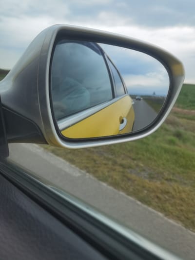 The image displays a passenger-side rearview mirror of a vehicle in transit near Zimnitsa, Bulgaria. The lower-left foreground is occupied by a dark gray interior door panel and the upper edge of the car's window. The central focus is a silver-framed, oblong side mirror.

The mirror's reflection depicts the rear quarter panel of the vehicle, which is yellow and includes a silver door handle. Beyond this, a straight, two-lane asphalt road recedes into the distance beneath an overcast sky. Two vehicles are visible on the reflected road; the closer one displays illuminated headlights. A dark, indistinct conical structure is present on the distant horizon. Green and brown grassy terrain flanks both sides of the road.

Directly outside the mirror's frame, the upper right portion of the image shows a cloudy sky. Below it, a grassy roadside embankment transitions to a gray road shoulder. Through the window, behind the mirror, a portion of the yellow car body and a light blue object within the vehicle are visible.
