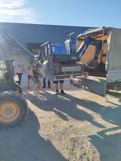 In Rozdil, Ukraine, a blue Scheppach mini-excavator is being loaded onto a flatbed trailer. The excavator is positioned on a wooden pallet, which is being supported by the trailer. A loader is attached to the front of the excavator, assisting with the loading process. Several individuals are present, observing or participating in the operation. The scene takes place outdoors on a dirt ground, with a large industrial building in the background and a clear blue sky above. The lighting suggests it is daytime.