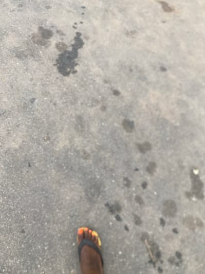 The image shows a close-up, blurry view of a person's right foot wearing a black flip-flop with brightly colored toenails (orange and yellow). The foot is positioned on a rough, grey asphalt surface that is speckled with dark, irregular splotches, possibly oil or water stains. The background is out of focus, but appears to be a continuation of the asphalt surface. The scene is not identifiable as being in Gulum, Nigeria.