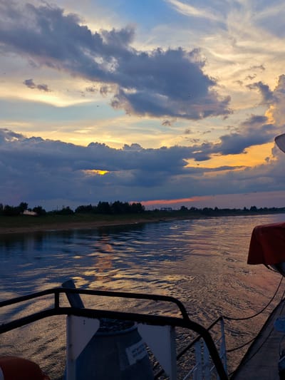 This media file depicts a scenic river view at sunset, likely from a moving boat. The foreground shows part of the boat's railing and deck, with a life preserver visible on the left and a white and blue cylindrical object with a label on the right. The water in front of the boat reflects the colors of the sunset, creating shimmering patterns.
The middle ground features a wide river with gentle waves. Along the riverbank, a line of trees and shrubs is visible, with a sandy or grassy shore. On the left bank, a white, boat-like structure rests on the shore, possibly beached or docked.
The sky is a dramatic blend of colors, characteristic of a sunset. Dark, heavy clouds are interspersed with patches of bright orange, yellow, and blue sky. The sun is not directly visible but its light breaks through a gap in the clouds, illuminating a portion of the sky with a golden glow. Several small birds are scattered across the sky.
The overall mood is tranquil and serene, enhanced by the warm hues of the sunset and the gentle movement of the water. The time of day is clearly sunset. The presence of a river, trees, and a distant shoreline suggests a rural or semi-rural setting. The location is contextualized as Vorogovo, Russia.
Visible text on the cylindrical object on the right reads "НАГНЕТАТЕЛЬ" and "ВЕНТИЛЯТОР" which translates to "PUMP" and "FAN" respectively.
