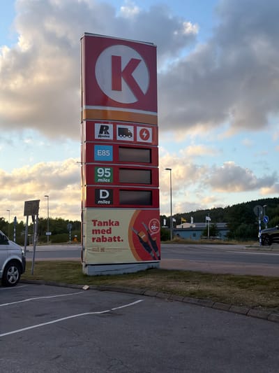 An outdoor medium shot shows a Circle K gas station sign with digital price displays. The sign features the prominent red Circle K logo at the top. Below the logo, there are several panels displaying fuel types and prices. From top to bottom, these include "R Routex" with icons for a truck and an electric car charger, "E85", "95 miles", and "D miles". A large banner at the bottom of the sign reads "Tanka med rabatt." (Fill up with discount.) and shows an offer with images of fuel nozzles. The sign is positioned on the right side of the frame, with a portion of a silver van visible on the left. The background consists of a road intersection, streetlights, traffic signs, a grassy area, and a treeline with a building and flags in the distance. The sky is partly cloudy with warm, late afternoon light. The scene appears to be located in Hogstorp, Sweden.