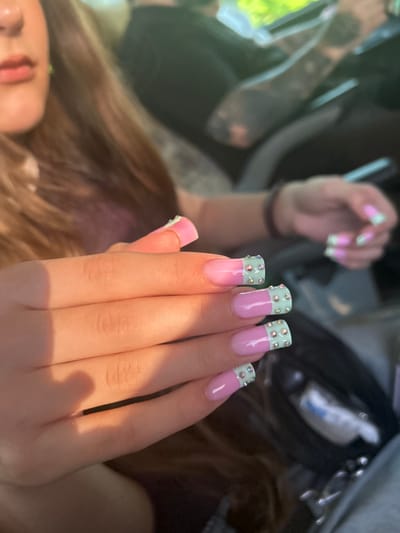 A close-up shot captures a woman's hands adorned with long, pastel pink and mint green French-tip acrylic nails decorated with small rhinestones. The woman, partially visible from the chest up, has brown hair and is seated inside a vehicle. The background shows a blurred view of another person, likely in the driver's seat, and the interior of a car. The lighting suggests it is daytime, possibly late afternoon, casting warm shadows on the woman's hands. There is no discernible information to place this image specifically in Maiaky, Ukraine.