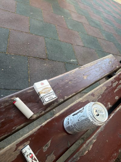 An outdoor scene on a weathered wooden bench, possibly in a park or public area. The bench sits on a paved surface made of interlocking dark and green rectangular tiles.
On the bench, from left to right, are three objects:
1. A white disposable lighter with a red button.
2. A pack of "Victory White" cigarettes, with a warning label stating "Smoking kills".
3. A partially consumed can of an energy drink, with a design featuring a stylized white pattern over a silver background. The can has text in what appears to be Cyrillic script.
The image is taken from a high angle, looking down at the bench and the items on it. The lighting suggests it is daytime. There are no people visible in the image, and no clear indication of the specific location beyond it being an outdoor, urban or semi-urban setting. There is no information in the image to suggest it is in the city of Chelopech, Bulgaria.