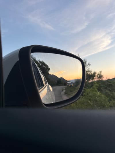 The image depicts a reflection in a car's side mirror, showing a winding road at dusk. The scene is set in the municipality of Svinjišta, Montenegro. A white car is visible in the mirror, with its headlights on, driving on the road. The sky is a blend of blue and orange, indicating the sunset. Green vegetation and trees line the sides of the road, and mountains are visible in the distance. The reflection also captures a portion of the car the photo is taken from, specifically its white exterior.