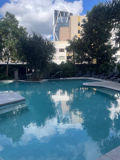 An outdoor swimming pool is shown in the foreground. The pool is filled with bright blue water and reflects the sky and surrounding environment. Around the pool are lounge chairs and lush green plants. In the background, there are several modern buildings. The scene appears to be in an urban setting, likely a hotel or residential complex. This image was taken in Brisbane, Australia.