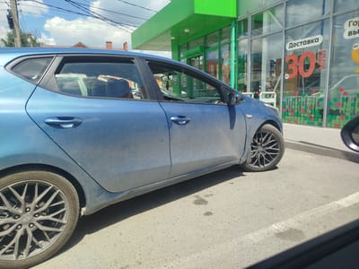 The image shows a blue Kia hatchback parked in front of a green building with large windows. The building has signage that includes the word "Доставка" (Delivery) and the number "30". The car is facing right, and its driver's side is visible. The sky is blue with some clouds, and power lines are visible overhead. The car's wheels are black with silver accents. The ground is asphalt, with some white markings. The scene appears to be a commercial area, possibly a store or a service center.