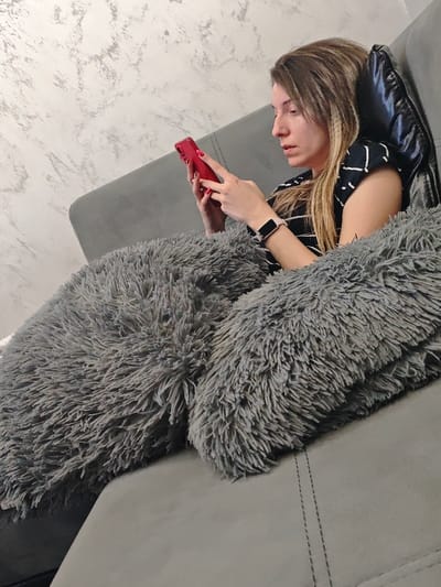 An adult female is depicted in a residential interior located in the city of Opaka, Bulgaria. She is seated on a gray fabric sofa, leaning back with her head resting against a dark cushion or the sofa back. Her hair is long, straight, and light brown. She wears a dark top with a white pattern and a light-banded smartwatch on her left wrist. Her fingernails are painted red. The female is actively engaged with a red-cased smartphone, holding it with both hands and directing her gaze downwards towards its screen. A large, shaggy, dark gray faux fur or plush blanket covers her lower body and portions of the sofa. The background consists of a light-colored wall with a textured, mottled gray pattern.