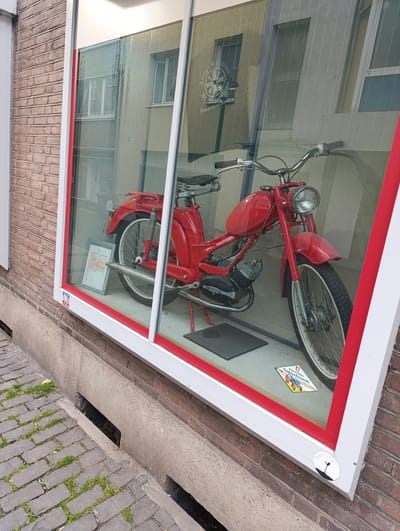 A red motorcycle is displayed in a storefront window, a sign with a logo reading "Jung-Schützen" is visible in the reflection on the glass. The brick building is located in Emmerich on the Rhine, Germany. A sticker reading "Emmericher Traktorfreunde" is on the glass. A stone pathway with grass is visible below the window. Another sticker reading "plantifol" is in the bottom corner of the window.