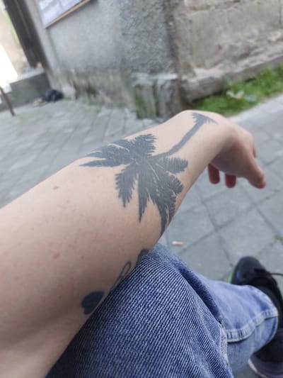 The image shows a close-up view of a person's forearm with a palm tree tattoo, resting on their lap wearing blue denim jeans. The background is blurred, suggesting an outdoor urban environment. Paved ground with square tiles is visible, along with a stone wall and some greenery. A discarded cigarette butt is present on the ground. The scene is likely located in Edineț, Moldova, based on the provided context.