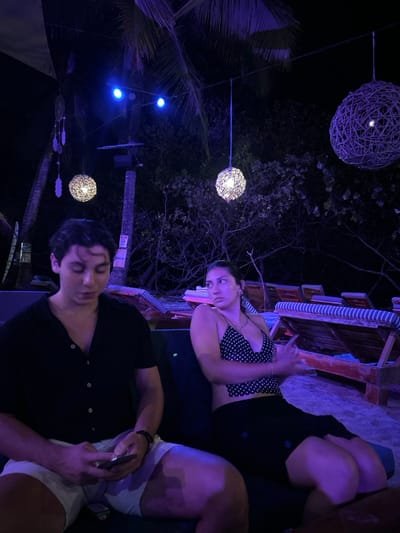 The scene is a dimly lit outdoor space at night, located at the coordinates 9.632, -85.158 in Santa Teresa, Costa Rica. Two individuals are seated together on a wooden bench. The male subject, who is dressed in a black button-down shirt and white shorts, is seated to the left, looking at his phone. The female subject is dressed in a black and white polka dot tank top and black skirt, seated to the right, looking away from the camera. She is sitting with her arms crossed, looking straight ahead. There are multiple wooden benches in the background, indicating an outdoor seating area. The area is illuminated by multiple hanging string lights. There is lush foliage overhead, and there are a few trees with branches that create a canopy of green leaves and branches, partially illuminated by the string lights. The scene is peaceful and tranquil, creating a quiet and relaxing atmosphere.