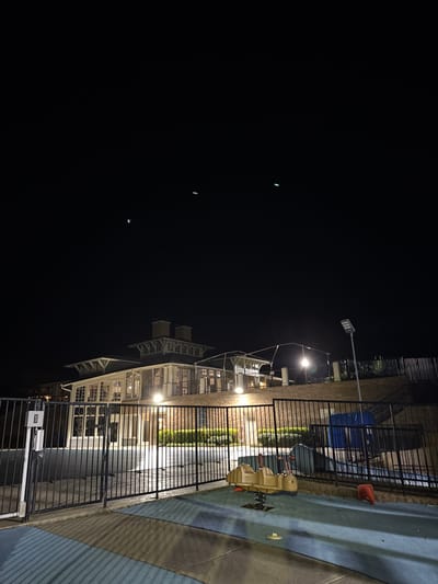 Night scene featuring a brightly lit two-story building with multiple windows and a distinct architectural style. A black metal fence runs along the perimeter, separating the building from a playground area with a spring rider toy. Streetlights illuminate the area, and a construction cone is visible in the playground. The sky is dark with a few distant light points.
