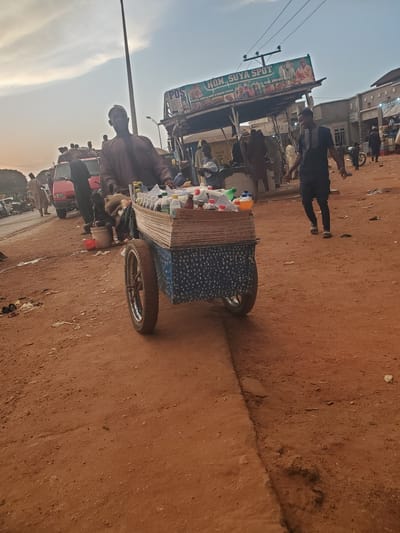 In Funtua, Nigeria, a street vendor pushes a cart laden with bottles of beverages. The cart is covered with a blue and white patterned cloth, and its wheels are partially visible. The vendor, dressed in traditional attire, is partially obscured by the cart, but his head and shoulders are visible. In the background, a red vehicle is parked, and several other people are seen walking around. A sign above a stall reads "HON. SUYA SPOT," with a phone number and address. The ground is made of dirt, and there is debris scattered around, suggesting a bustling market environment. The sky is overcast with a hint of sunset.