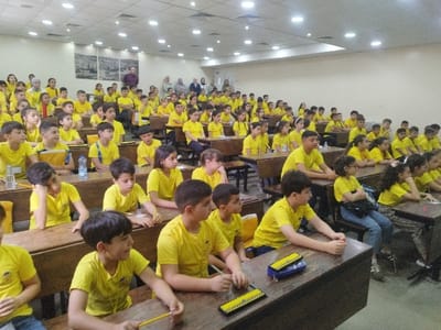 The image displays an indoor scene within a tiered lecture hall or large classroom located in the city of Al-Khalil, Palestinian Territory. Approximately 100 to 120 children, predominantly between 7 and 14 years of age, are seated in rows of dark wooden desks and benches. Nearly all children are uniformly dressed in bright yellow t-shirts. They are observed in attentive postures, mostly facing forward, with some individuals looking towards the sides. Items visible on the desks include pencils, paper, blue abacus tools, and several water bottles. In the background, at least ten adults are present, either standing or seated at the rear of the hall. The room features light-colored walls, an acoustic tile ceiling equipped with numerous recessed lights and some linear fluorescent fixtures, and two framed black-and-white images depicting historical or architectural scenes mounted on a wall section at the back. The environment is brightly and evenly illuminated.