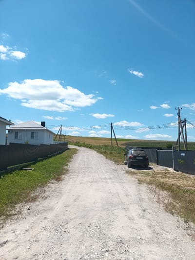 The image shows a dirt road leading towards a rural residential area under a clear blue sky with scattered white clouds. On the left, a white house with a metal roof and dark window frames is visible, bordered by a dark metal fence. To the right of the road, a dark-colored car is parked near a dark metal gate and fence. In the background, a field of dry grass stretches to the horizon, with utility poles and power lines crossing the scene. The overall environment suggests a sunny day in a suburban or rural setting.