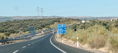 The image shows a highway scene near Almaraz, Spain. A paved road curves to the right, with a metal guardrail on the left. The surrounding landscape is arid and hilly, covered with low-lying green shrubs and some taller, dry bushes. In the distance, two large metal pylons stand against a clear blue sky. A blue highway signpost stands on the right side of the road, indicating directions and distances to various locations, including "700 m," "6.4 km," "24 h," and "21 h." There is some smoke visible rising from the left side of the highway in the mid-ground. Several vehicles are present on the road, including at least one white car.