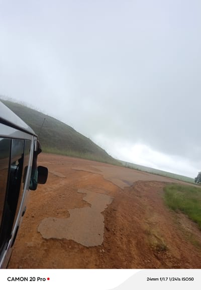 An over-the-shoulder, low-angle perspective from inside a moving vehicle shows a person's arm and hand protruding from the passenger window, reaching upwards as the vehicle travels on a dirt road. The road is a reddish-brown color, with patches of broken asphalt or concrete. To the left of the road, a grassy hill slopes upward, covered in sparse vegetation and shrouded in a light mist or fog. The sky is overcast and white. The vehicle's side and window are visible on the left. The image appears to be taken in Nguroje, Nigeria, as indicated by the text overlay, though no specific landmarks are identifiable within the scene.