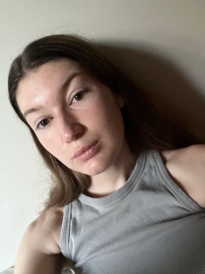 The media file is a portrait of a young person. The subject is a person with long, brown hair, looking towards the camera with a neutral expression. They are wearing a grey ribbed tank top. The background is a plain wall, with a shadow cast on it, suggesting an indoor setting. The lighting is soft, with some highlights on the subject's face, indicating it might be taken in the evening or in a room with artificial light. There is no visible text in the image. The location context provided is Skawina, Poland.