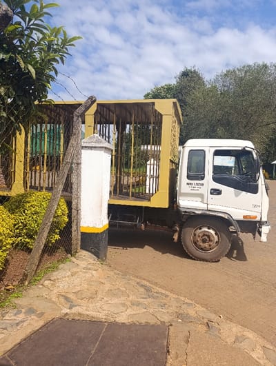 A white, medium-duty truck, possibly an Isuzu, with a distinctive yellow, open-caged structure mounted on its bed, is parked next to a fenced entry point in Kaplelwo, Kenya. The truck's cab is white with black trim, and its driver's side door displays visible text: "MPPUYA FACTORY" followed by "P. O. BOX 600 - 412". The truck's wheels show signs of use, consistent with an active working vehicle.
To the left of the truck, a sturdy concrete fence post, painted white with a black and yellow base, marks the entrance. A chain-link fence, topped with barbed wire, runs behind the post and along the left side of the frame. Lush green foliage, including bushes and a tree, grows densely behind and around the fence. The foreground features a reddish-brown ground surface transitioning from paved cobblestones or flagstones to a dirt path, with a dark metal manhole cover partially visible at the bottom.
Beyond the fence and truck, a backdrop of numerous green trees extends towards the horizon, under a bright blue sky dotted with scattered white clouds, indicating clear, sunny daytime conditions. No people are visible in the scene, and the truck appears stationary, suggesting it is either waiting, having arrived, or about to depart from a facility. The caged bed of the truck hints at specialized transportation, possibly for livestock or specific cargo related to the "MPPUYA FACTORY." The overall setting appears to be an outdoor, semi-rural, or industrial area.