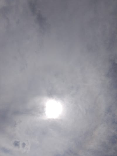 The image shows a sky with clouds and the sun partially obscured. The sun appears as a bright, white circle with rays emanating outwards, surrounded by a hazy glow. The clouds are light gray and wispy, with some darker patches indicating denser formations. The overall impression is that of a partly cloudy day. There is no indication of the city of Aqköl, Kazakhstan, or any specific urban environment.