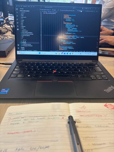 This image captures a close-up of a black Lenovo ThinkPad laptop, its screen displaying code in a dark theme editor. The laptop is placed on a wooden desk, alongside an open notebook filled with handwritten notes in red ink. A black pen rests on the notebook, partially obscuring the writing.

The screen shows a file explorer pane on the left, with a directory structure visible, and the main area displays lines of Python code. The file `catalog.py` seems to be open, with several lines of code related to grid column definitions and dashboard options. The status bar at the bottom of the editor indicates the time as "18:57" and the date as "24/06/2026".

The notebook has handwritten text, including dates and what appear to be tasks or project notes. Some words are underlined, and there are small red sketches. One entry, dated "25/06/", mentions "UPLOAD PRES THALAMENTATION PLAN" and "MORITION TEST CON ACIENTE POMENICO". Another entry reads "WORKING STATUS."

The setting appears to be an indoor workspace, possibly an office or home office, given the laptop and notebook. The lighting suggests it is daytime, though the indoor environment makes it difficult to determine the time of day definitively beyond the digital clock on the laptop. The overall mood is one of focused work and planning. There are no people clearly visible, though hands can be seen in the upper left corner, suggesting someone is using the laptop. The presence of the Intel Core i7 sticker on the laptop indicates a powerful processing unit. The location context provided is Basiglio, Italy.
