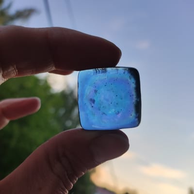 A close-up, low-angle shot shows two human fingers holding a blue, square-shaped glass object against a backdrop of a cloudy sky and blurred green foliage. The glass object has a slightly iridescent quality, with darker blue swirls and small dark specks embedded within it. The fingers are positioned at the top and bottom of the glass, obscuring parts of it. The skin of the fingers appears textured. The lighting suggests it is either sunrise or sunset, with soft light illuminating the scene. There are no discernible landmarks or contextual cues in the image that would allow for identification of the location in Kineshma, Russia.