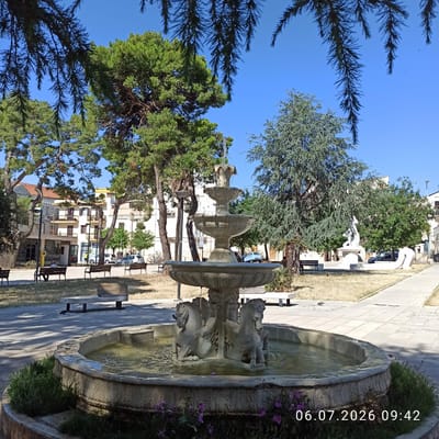 This is a medium shot of a tiered fountain in a park on a sunny day, located in Ruvo di Puglia, Italy. The fountain is made of stone and features sculpted horses at its base. Water cascades from the top tier to the lower basins, creating a gentle spray. The park has paved walkways, benches, and well-maintained grassy areas. Mature trees, including pine and cedar varieties, provide shade. In the background, buildings with apartment units are visible, suggesting an urban setting. The sky is clear and blue, indicative of pleasant weather. A timestamp in the lower right corner reads "06.07.2026 09:42". There are no people clearly visible in the foreground, but the presence of benches suggests it is a public space. The overall scene depicts a tranquil park environment.