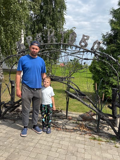 In Breytovo, Russia, a man and a young boy stand side-by-side outdoors. The man, wearing a blue t-shirt, grey sweatpants, and blue Crocs, is on the left. To his right stands the boy, in a white t-shirt and patterned pants, wearing blue and grey Crocs. They are positioned in front of an arched, ornate metal structure with wire-wrapped branches and what appear to be decorative letters spelling out a word, partially obscured by foliage. Behind them, a lush green lawn stretches towards trees and a cloudy sky. The ground in the foreground is paved with grey brick. The overall scene suggests a casual outdoor setting, possibly a park or a decorative entrance to a property.