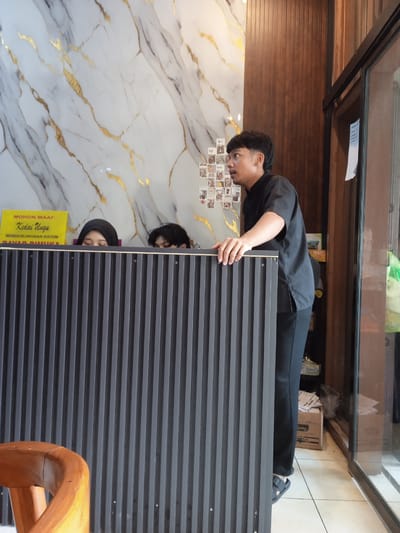 The photograph depicts an interior scene in Pandean, Indonesia. Two individuals are visible behind a gray, vertically striped counter. One individual wears a black hijab, while only the hair of the other is visible. A yellow sign with Indonesian text is above the counter, reading "Mohon Maaf Kedai Ungu MEMBERLAKUKAN SISTEM". A man in a black uniform stands beside the counter, resting his hand on its edge and looking to the right. The wall behind features a decorative marble pattern with gold veins and a display of small photographs. A wooden wall panel and a glass door are to the right, revealing a glimpse of the exterior. A wooden chair is partially visible in the lower left corner.