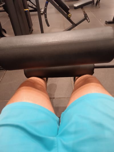 A first-person perspective from a seated position shows a leg extension machine at a gym. The machine's padded bar rests on the lower legs, just above the ankles. Light blue shorts are visible, covering the upper thighs. The floor is tiled in dark gray, contrasting with the light-colored skin on the legs. Gym equipment is blurred in the background, suggesting a shallow depth of field.