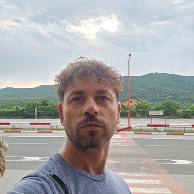A man with curly brown hair and a beard stands in the foreground, taking a selfie. He is wearing a blue t-shirt. Behind him, a road with white and red barriers is visible, with a pedestrian crossing marked by red and white stripes. In the background, a lush green mountain looms under a cloudy sky with the sun partially visible through the clouds. A street lamp with a red pole is also present in the background. A building with a brown roof and green trees surround it is visible on the slope of the mountain. A sign with text in an unfamiliar alphabet is mounted on a fence. The scene appears to be in Zlatna Panega, Bulgaria.