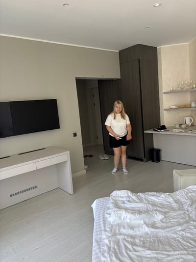 The image depicts an indoor scene, likely a hotel room, with a woman standing in the center. The room has light-colored wooden flooring and neutral-toned walls. To the left, a white desk with a black television mounted above it is visible. To the right, a large dark wood wardrobe stands against the wall, and next to it, a counter with shelves displays glassware and other items. A bed with rumpled white bedding is in the foreground, partially occupying the right side of the frame. The woman, wearing a white t-shirt and black shorts with white socks, is holding a white object in her right hand and appears to be looking towards the camera. The overall impression is a contemporary, clean, and possibly recently occupied living space. The specific location of Мядзельскі сельскі Савет, Belarus, is not directly discernible from the visual elements of the image.