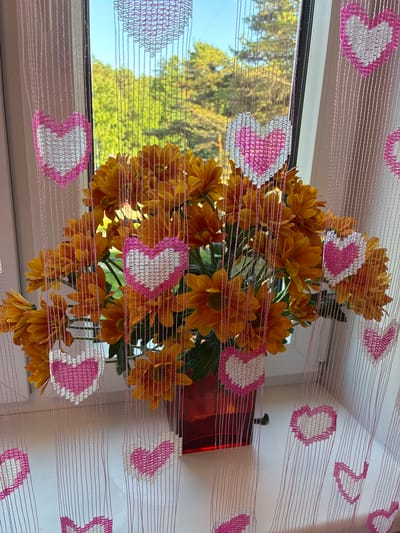 An arrangement of orange chrysanthemums in a red glass vase sits on a windowsill. The vase is partially obscured by a pink string curtain adorned with crocheted white and pink hearts. The window behind the flowers looks out onto a view of green trees and a blue sky.