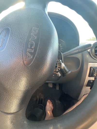 An interior shot of a car's dashboard and steering wheel, taken from the driver's perspective. A bare foot is visible, resting on the floor mat near the pedals. The car's ignition appears to be damaged or removed. The brand "DACIA" is embossed on the steering wheel. The scene is illuminated by natural light, suggesting it is daytime. The location is specified as Telenești, Moldova.