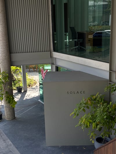 An exterior view shows a modern building with a large window and a sign reading "SOLACE." The building features metal siding, and a concrete awning hangs above the entrance. Lush green plants are present on the side. A view of a patio with furniture is visible through the awning. The ground is paved, and the image appears to be taken from an angle.