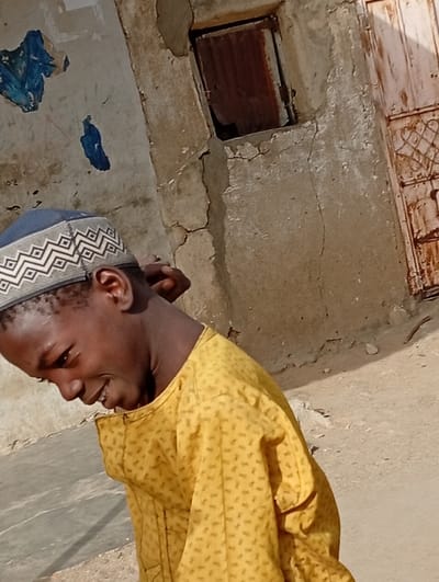 The scene, set in Gayawa, Nigeria, depicts a young male subject outdoors under direct sunlight. The individual has dark skin and is wearing a blue and white geometric-patterned kufi and a yellow long-sleeved garment with a subtle repeating pattern. His head is tilted downwards and to his left, revealing a smiling expression. His right hand is positioned behind his head. The background features a weathered, light-colored building wall exhibiting significant cracks and peeling paint. In the upper-right section of the wall, a small, square window opening is covered by a piece of rusted corrugated metal. To the far right, a portion of a heavily rusted door with embossed decorative elements is visible. The ground surface is dry, consistent with packed earth or sand.