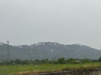 The image shows a landscape with mountains in the background and a telecommunications tower on the left. The sky is overcast, and there are water droplets on the camera lens, suggesting it is raining or has recently rained. In the foreground, there are green fields and trees. The scene appears to be in a rural or semi-rural area. Without further information, it is not possible to confirm or deny the location in the city of ნიგოითის თემი, Georgia.