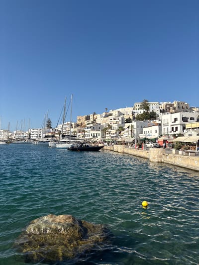 The image depicts the main harbor and waterfront of Naxos Chora, Naxos Island, Greece, viewed from the water. The foreground features calm, teal-blue water with a large, dark, partially submerged rock on the lower left and a bright yellow spherical buoy floating in the mid-right. Along the right, a light-colored stone quay functions as a pedestrian promenade, lined with various moored vessels including multiple white sailboats with visible masts and rigging, and a dark-colored rigid inflatable boat. Indistinct human figures are visible walking on the promenade and seated at outdoor tables of businesses fronting the harbor. A white pickup truck and other vehicles are parked on a street behind the promenade. The background is dominated by the densely packed white and light-colored Cycladic buildings of Naxos Chora, climbing up a hillside. Scattered green trees and shrubs are interspersed among the structures, with the elevated, more weathered buildings of the historic Kastro (castle) visible towards the top left of the hillside. The sky above is clear and deep blue, indicating bright, sunny conditions.