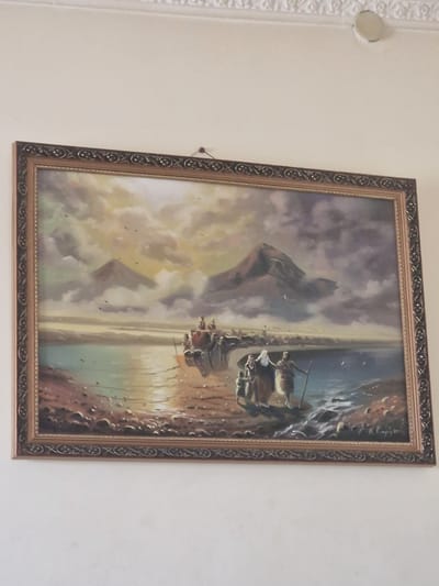 A framed painting hangs on a light-colored wall. The painting depicts a serene landscape with a body of water in the foreground and mountains in the background under a cloudy sky. Several figures are visible, including a group of people standing on the shore, appearing to be a family or a small procession, with one individual holding a staff. In the water, a small boat or raft is present, carrying a few figures. The overall atmosphere of the painting is peaceful and evokes a sense of journey or contemplation. The painting is framed with an ornate, dark-colored frame with gold accents. The wall itself shows some texture and appears to be part of an indoor setting, possibly a home or a gallery in Nor Kharberd, Armenia.