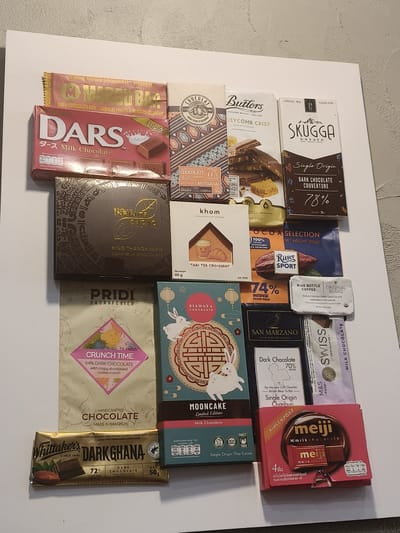 The image displays a collection of various chocolate bars and boxes arranged on a white surface. The chocolates are diverse in brand, origin, and type, including dark chocolate, milk chocolate, and some with added ingredients like fruits, nuts, and shortbread. Brands visible include Marou Bar, Dars, Infinite Cacao, Pridi Cacao Fevrier, Whittaker's Dark Ghana, Butlers, Skugga, Khow, Ritter Sport, Blue Bottle Coffee, San Marzano, Swiss, and Meiji. Some packaging features details about the cacao origin, percentage of cocoa, and manufacturing information. The arrangement suggests a display or collection of these confectionery items. There is no indication of the location being Chiang Rai City Municipality, Thailand.