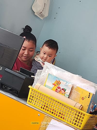 A woman is holding a baby in front of a computer in what appears to be a store or office. The woman, with her hair in a bun, is wearing a red shirt and looking down at the computer. The baby, wearing a striped shirt and a bib, is looking towards the right side of the frame. Behind them, a blue wall is visible, and a white plastic bag with Vietnamese text that appears to say "ANH TUYET STORE" hangs from the wall. In the foreground, a yellow plastic basket is filled with various items, including rolled-up papers, books, and what looks like a blue pen. The scene is indoors, with artificial lighting. The lower right corner of the image shows a "realme" watermark and the text "Shot by zsm_travel 2026.06.24 07:41", indicating the photo was taken on June 24, 2026, at 7:41 AM. The overall impression is a casual moment captured between a caregiver and a child in a working environment in Gò Dầu, Vietnam.